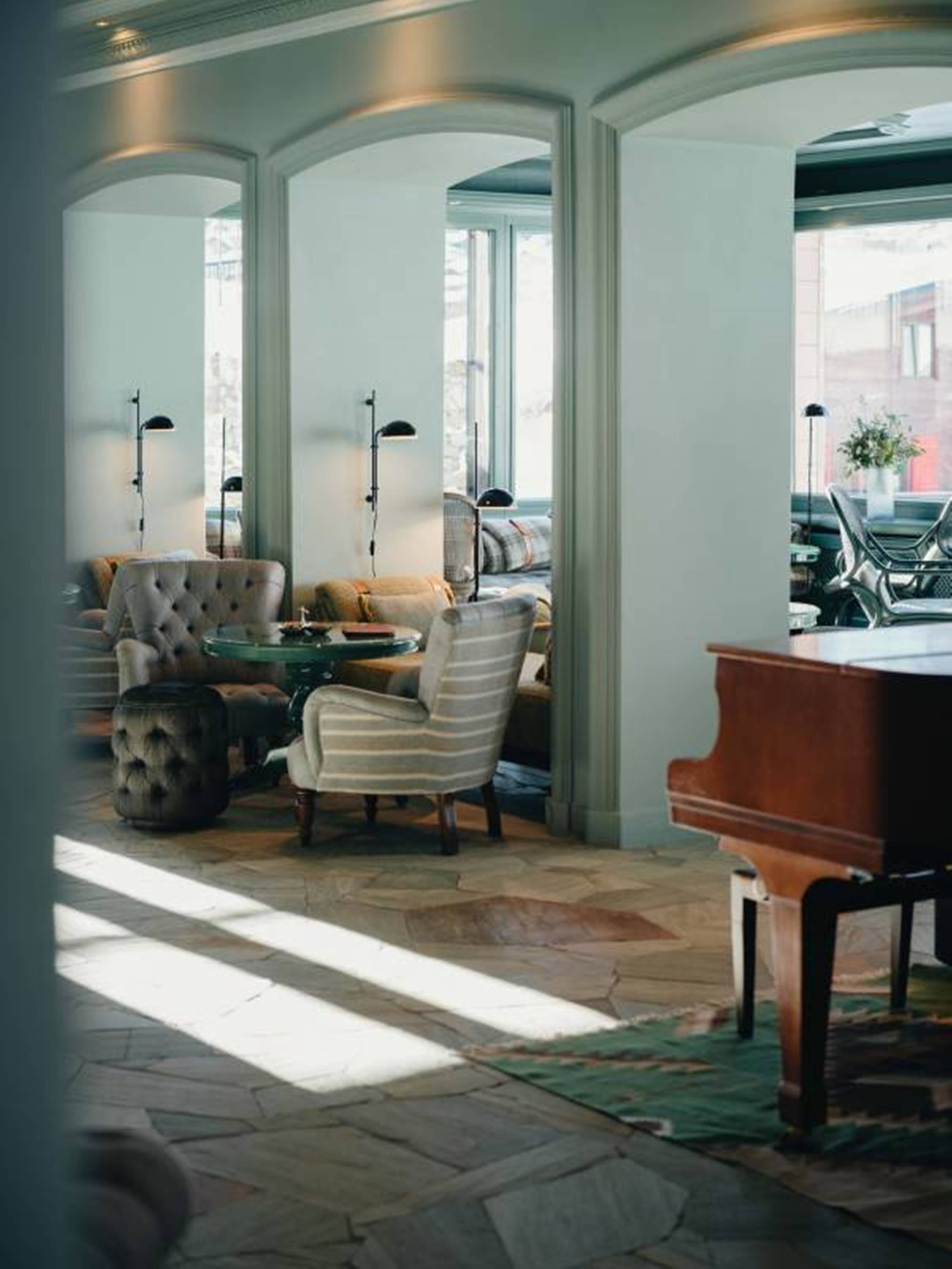 A room with a piano, a striped armchair, a tufted armchair, and a sofa. There are three floor lamps and a window with a view of the street.
