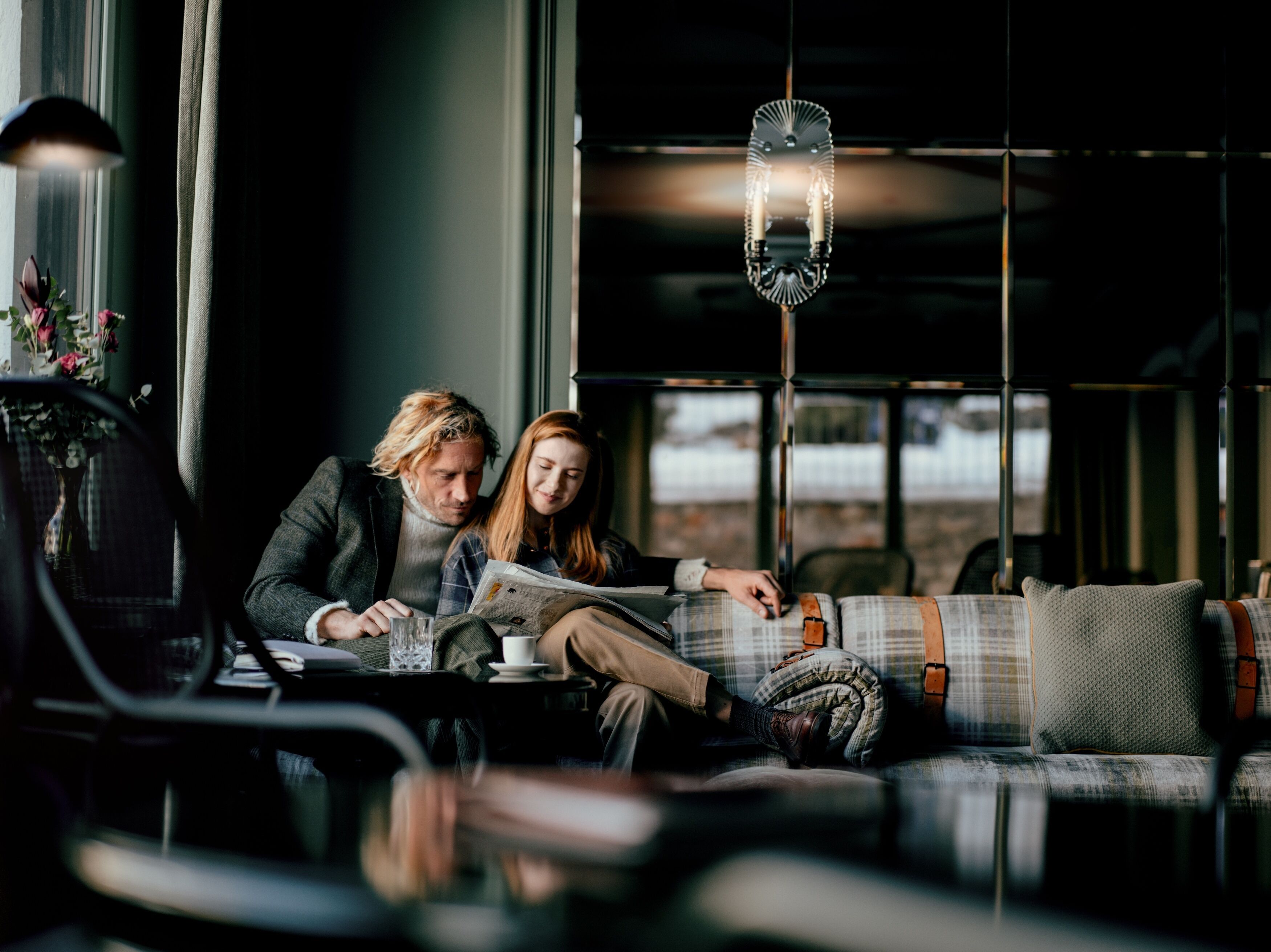 A couple sitting on a plaid sofa with a city view, reading a newspaper.