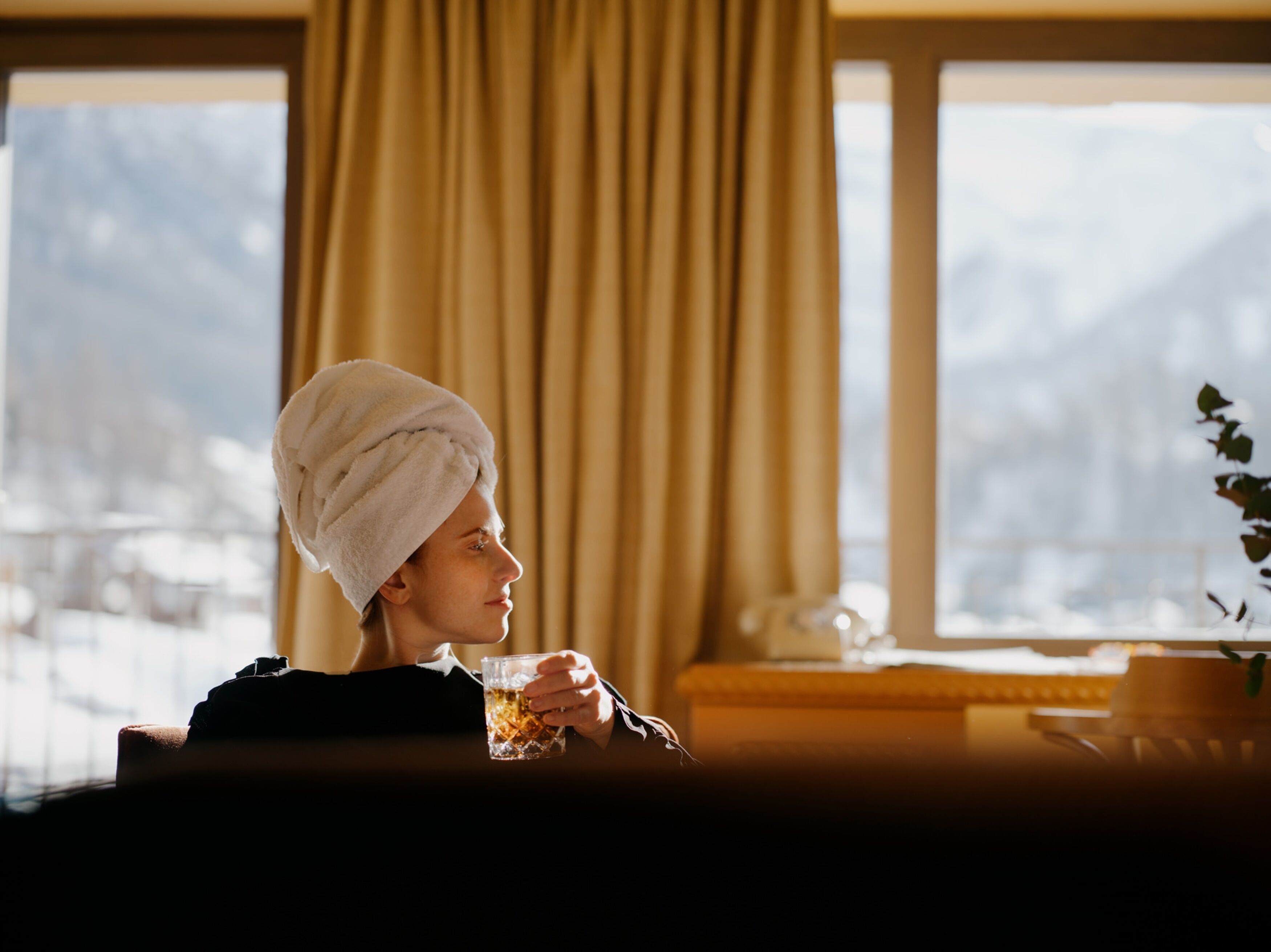 Une personne assise près d’une fenêtre, avec des rideaux, une serviette sur la tête et un verre contenant une boisson dans la main.