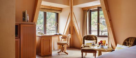 Un dormitorio acogedor en un ático con techo de madera, un ventanal con vistas a un pueblo, una silla cómoda, una mesita con una taza y una cama.