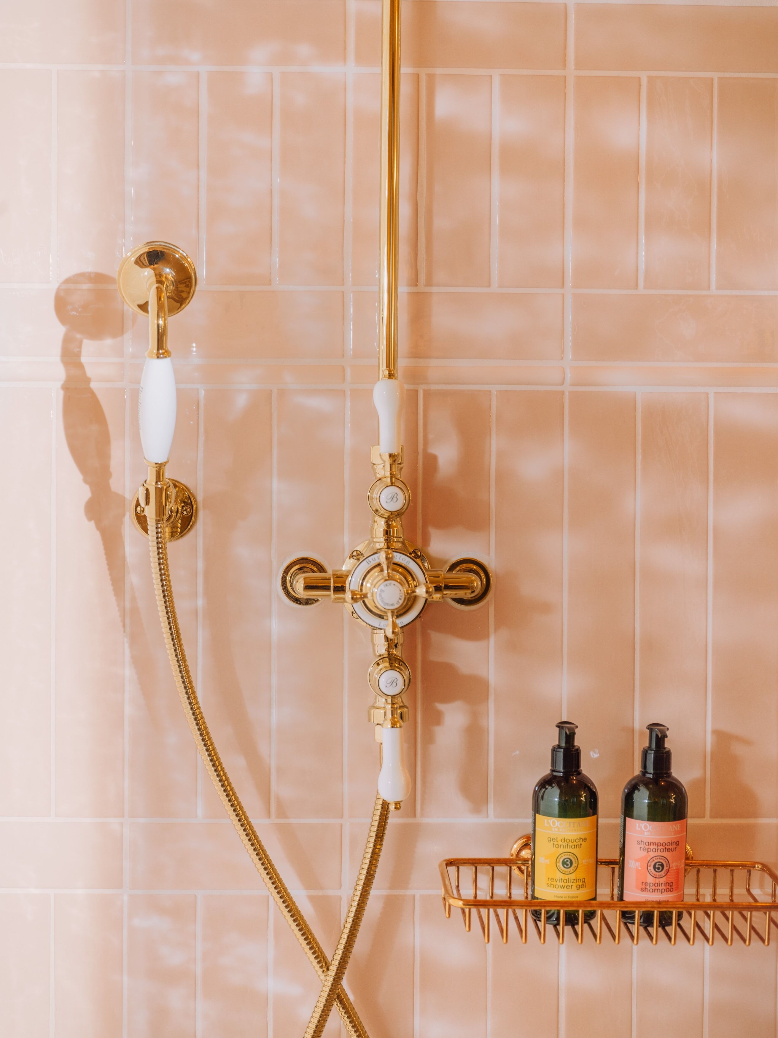 A bathroom with a gold shower system, a white handheld showerhead, and two bottles of L'Oreal products on a gold shelf.