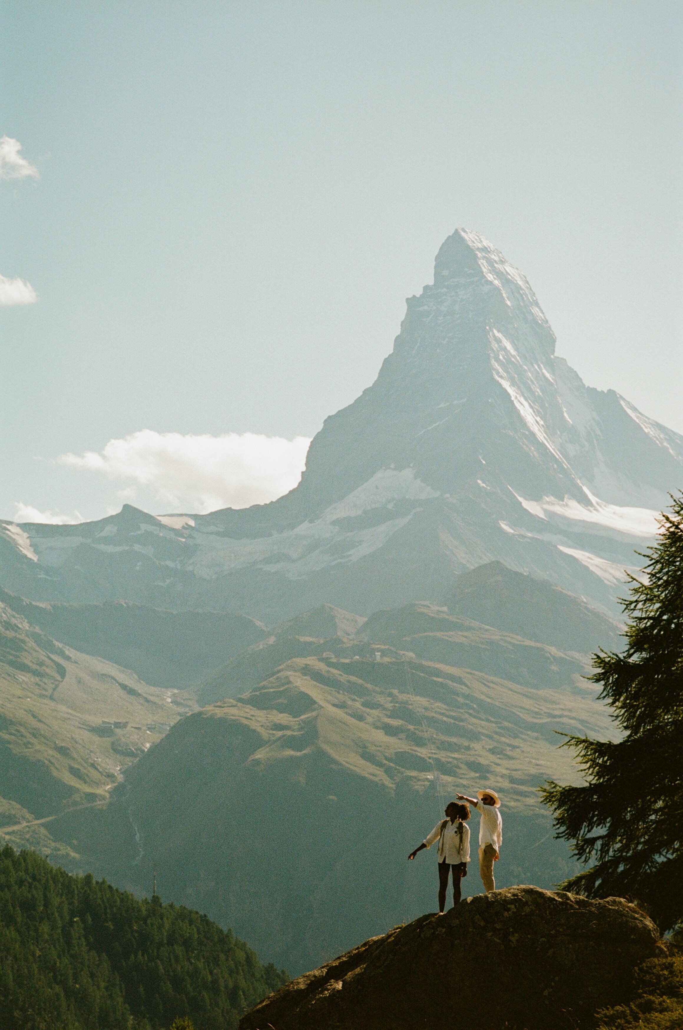 A mountain landscape with a prominent peak, likely a famous mountain, and two people standing on a rocky outcrop.