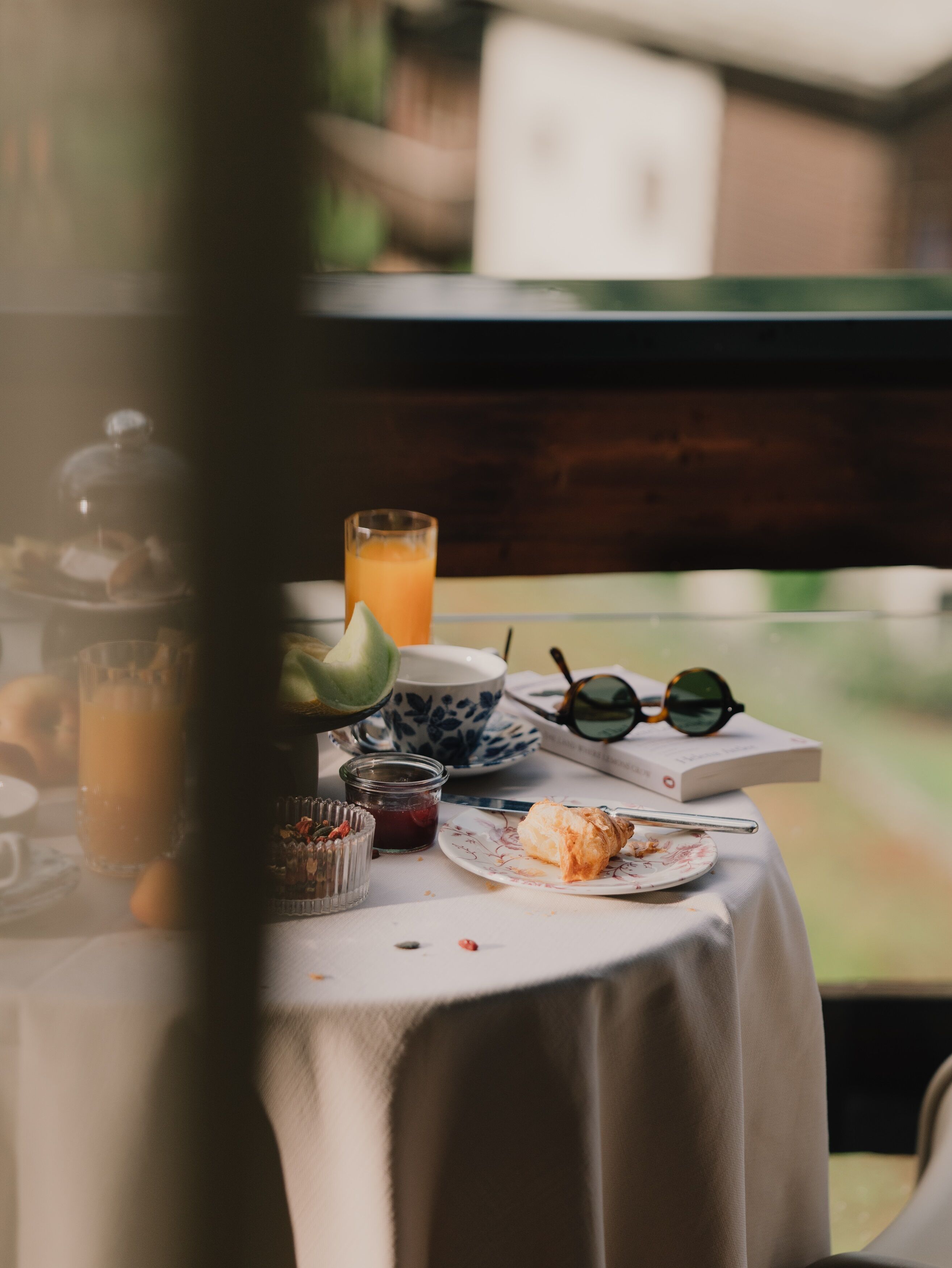A table with a glass of orange juice, a plate with a pastry, a jar of jam, a pair of sunglasses, and a book.