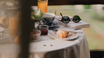 A table with a glass of orange juice, a plate with a pastry, a jar of jam, a pair of sunglasses, and a book.