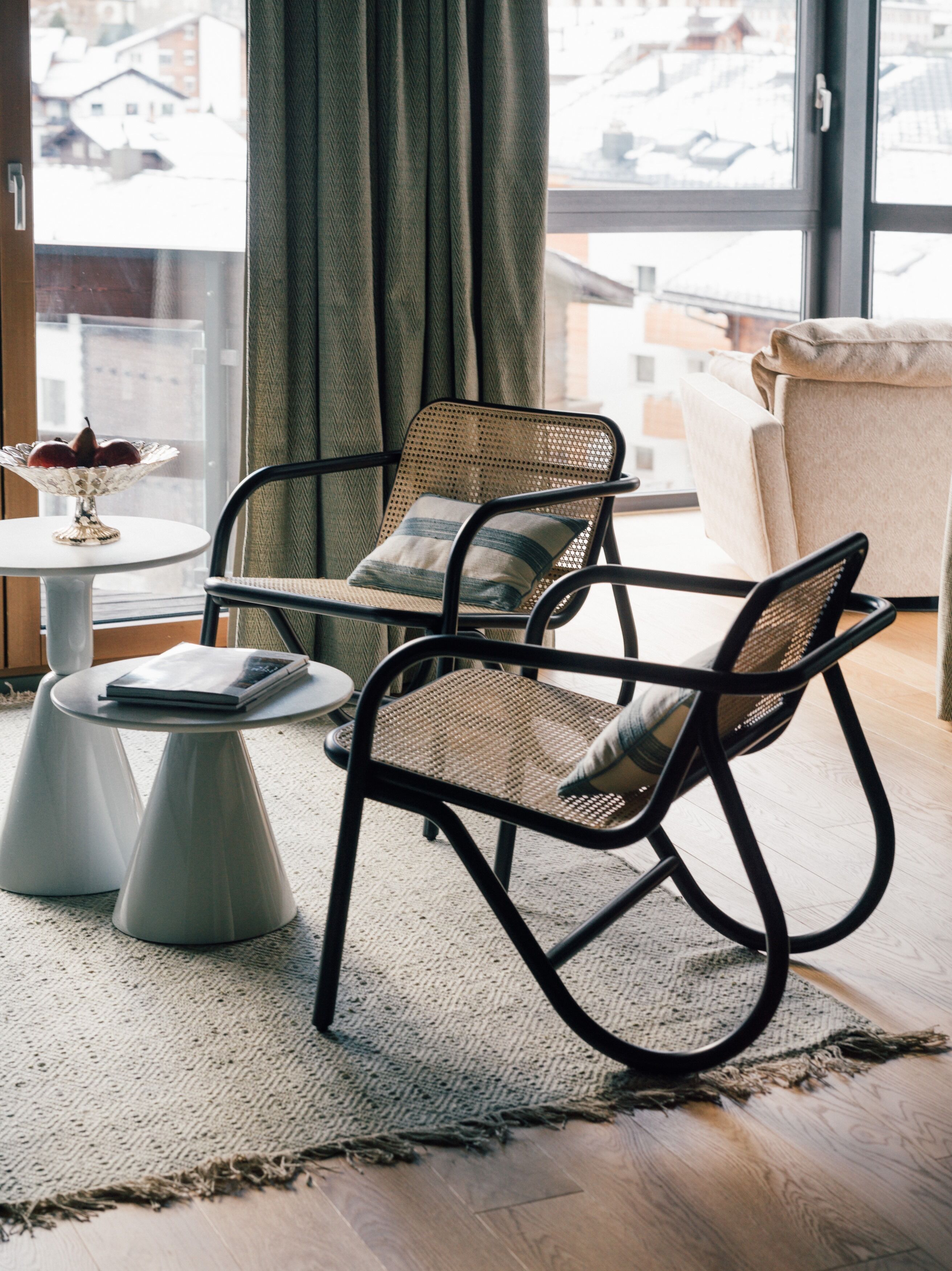 A modern living room with a wicker chair, a small round table, and a view of the city through large windows.