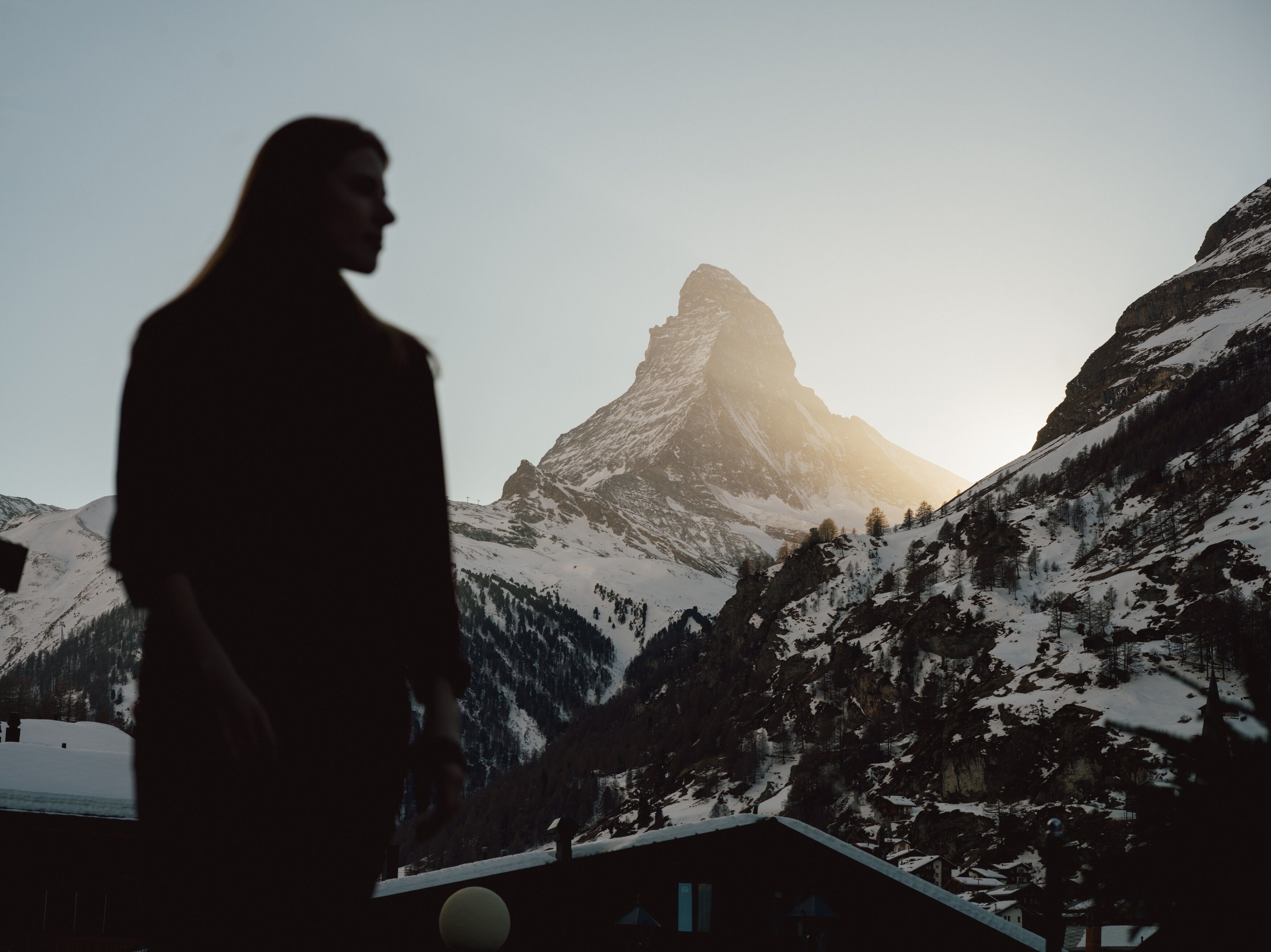 A person standing in front of a mountain with a snow-covered peak, overlooking a valley with trees and buildings.