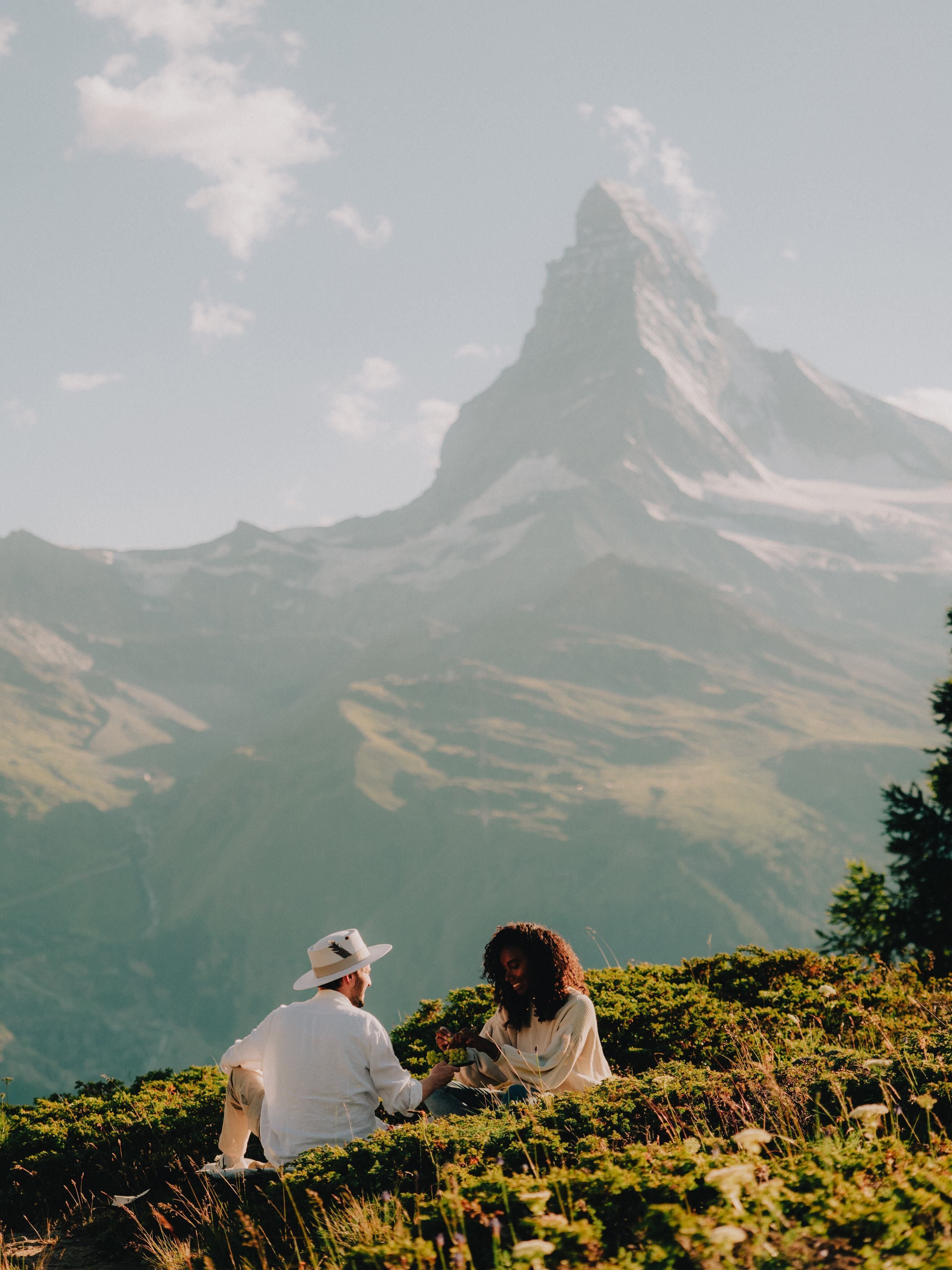 Two people sitting on a grassy hill with a mountain in the background.