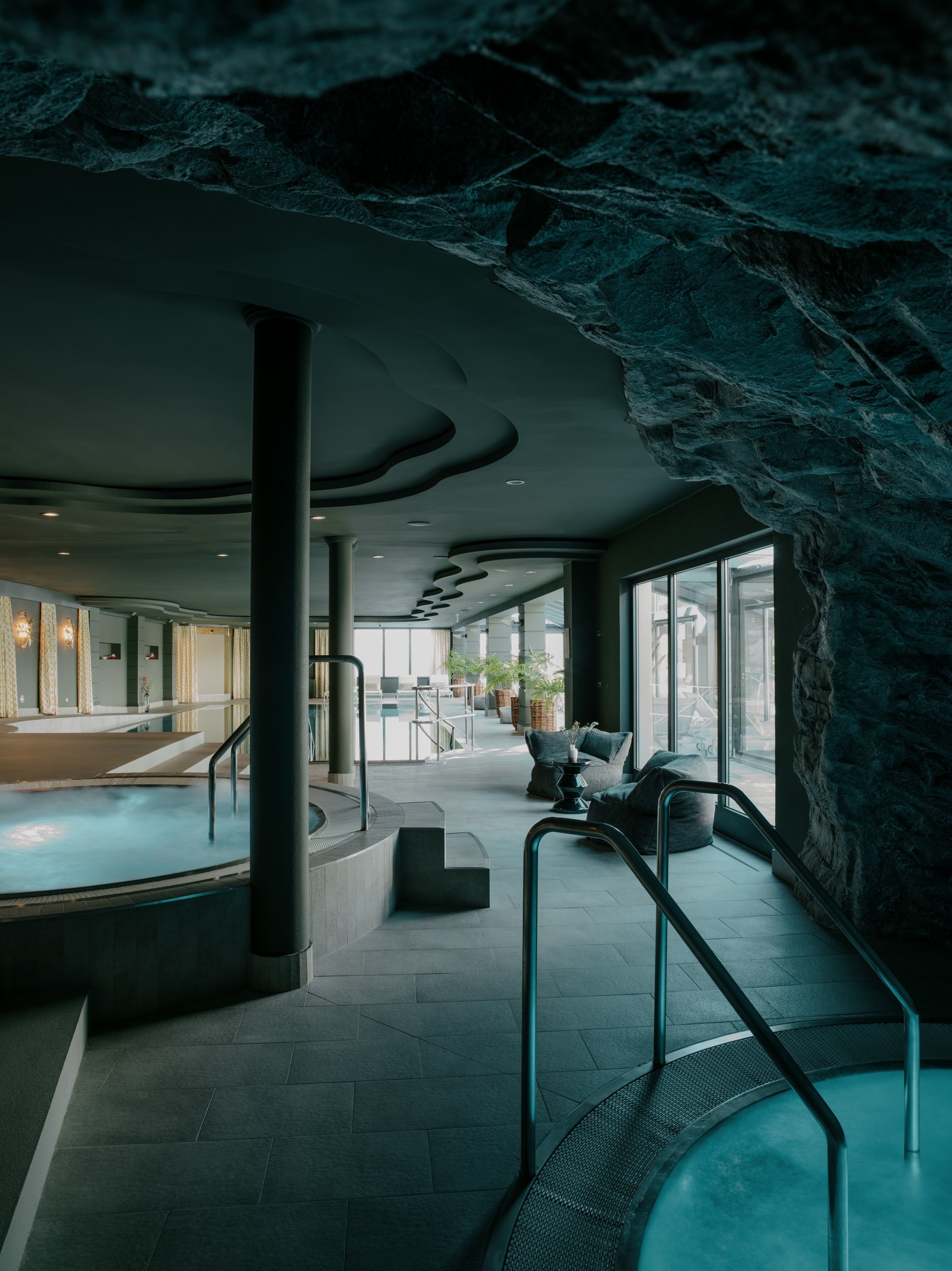 A modern indoor pool area with a unique rock-like ceiling, comfortable seating, and a view of the outside.