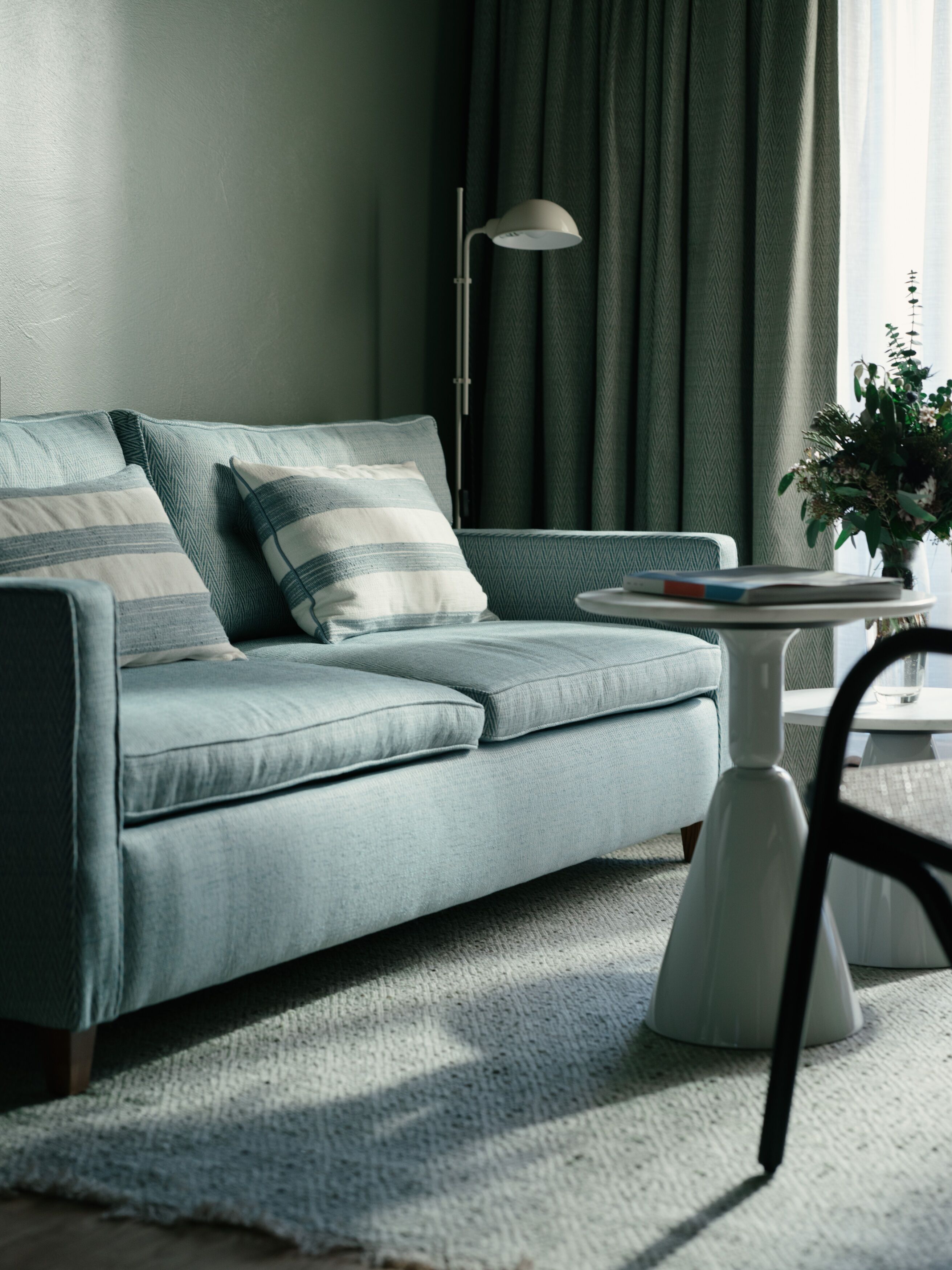A modern living room with a light blue sofa, a white side table, a floor lamp, and a green curtain.