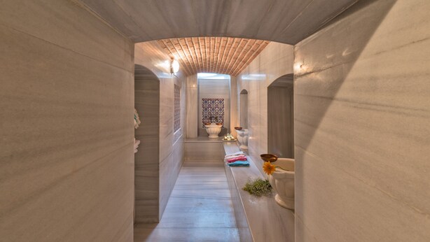 Un estrecho pasillo con baldosas, techo de madera y vista a una habitación con pared de azulejos y una puerta blanca.
