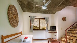 Un dormitorio con cama, ventilador de techo, televisor sobre una estantería, ventana con cortinas y una escalera con pared de piedra.