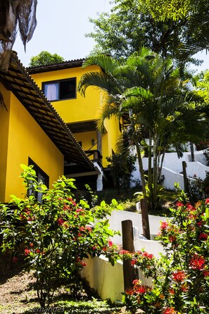 A yellow building with a balcony, surrounded by lush greenery and flowering plants.