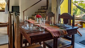 Una mesa de comedor de madera dispuesta con cristalería y un florero con flores rojas, con una escalera de piedra y una vista de casas al fondo.
