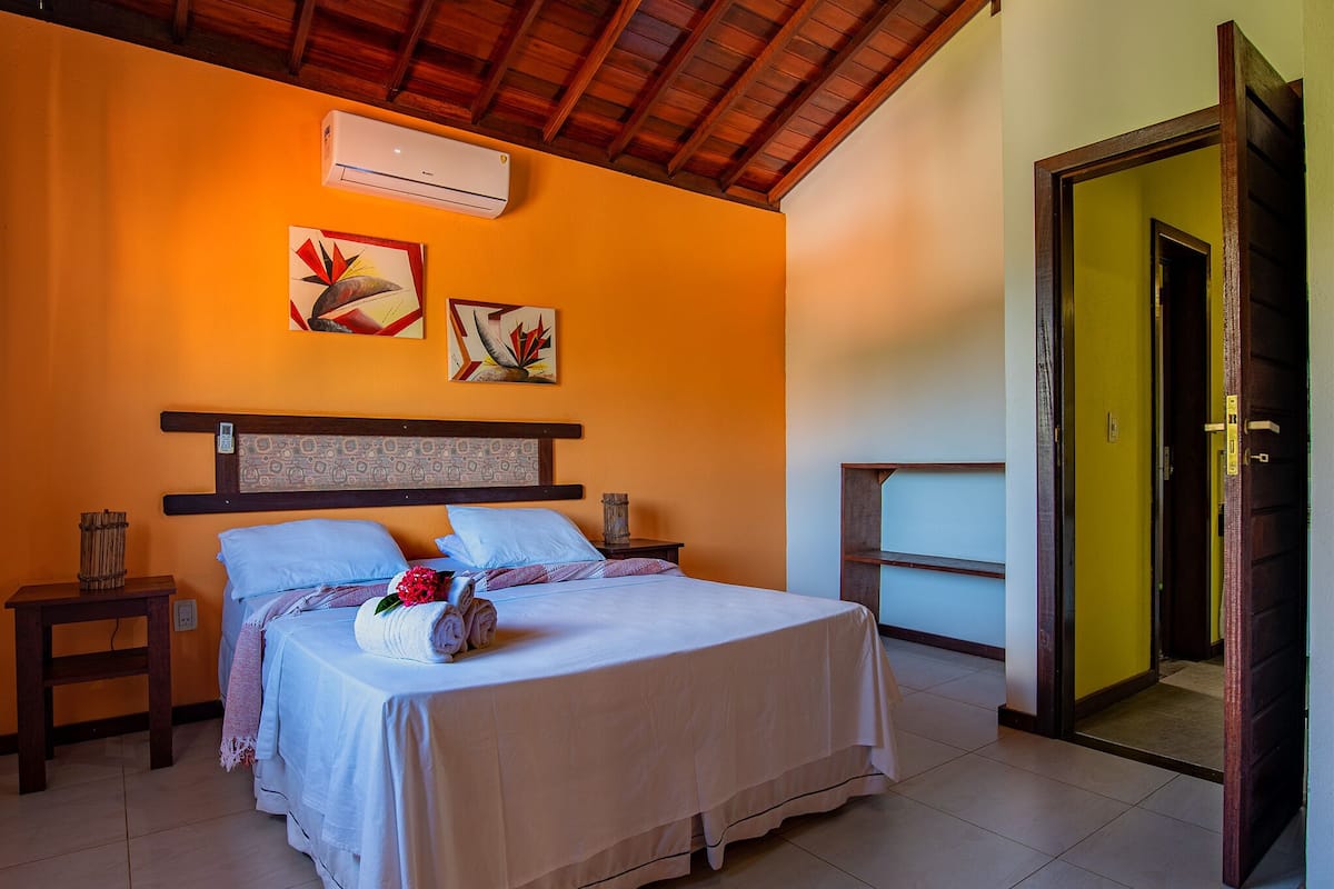 A hotel room with a bed, a nightstand, an air conditioning unit, and artwork on the wall.