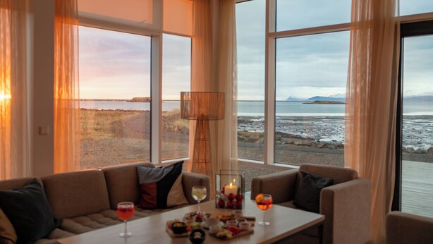 A room with a large window overlooking a beach, a sofa, a table with drinks and snacks, and a lamp. - Höfn - Berjaya Iceland Hotels (Höfn)