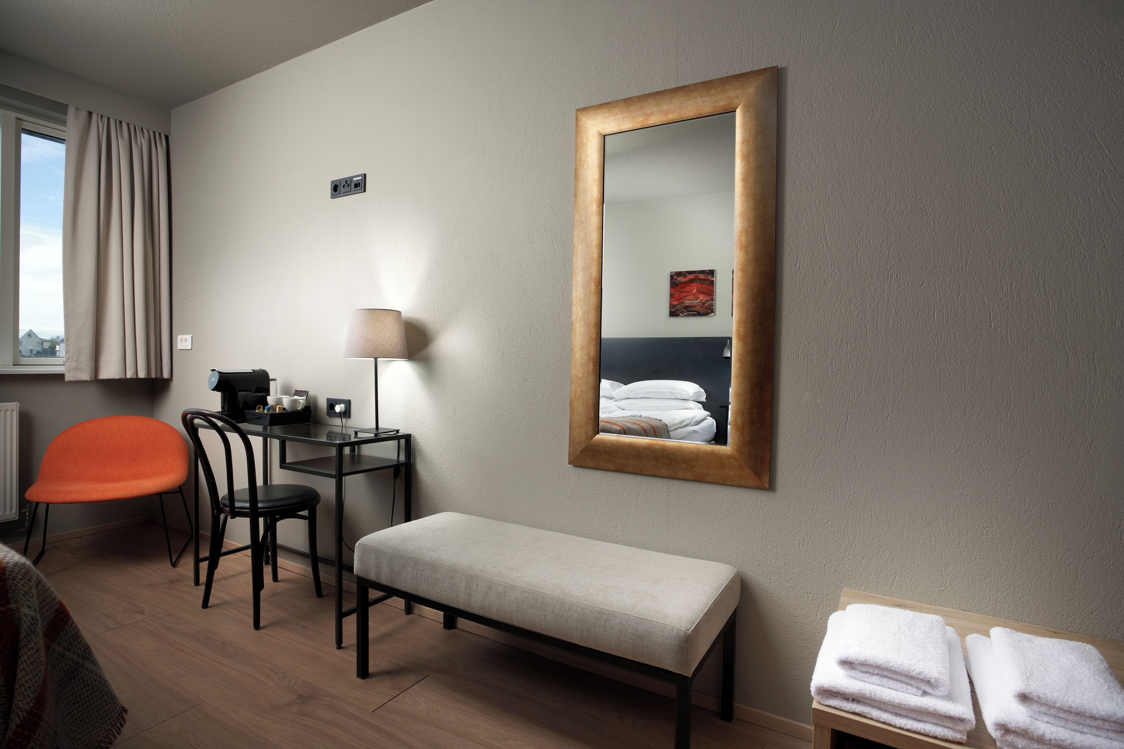 A hotel room with a bed, a desk with a lamp, a chair, a bench, and a mirror on the wall.
