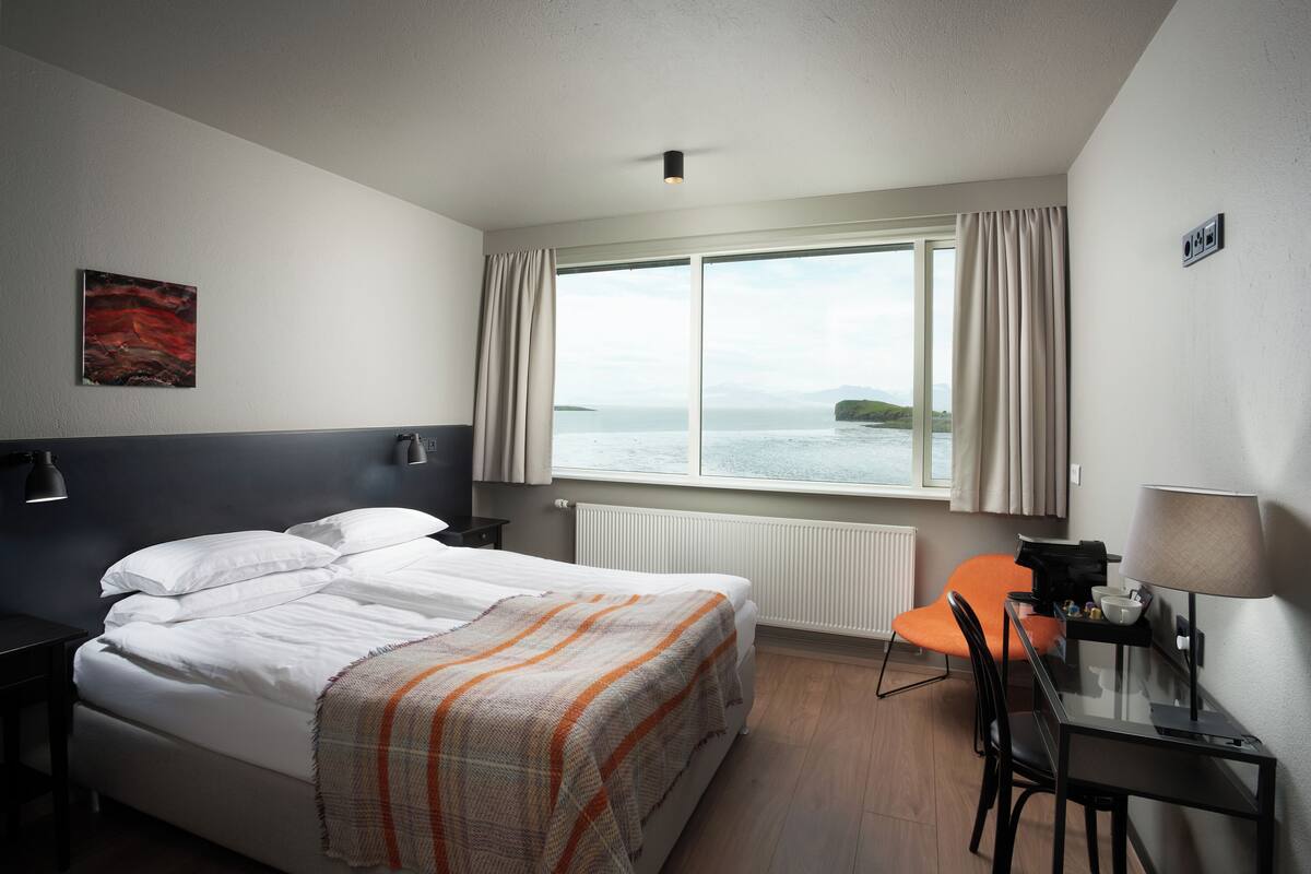 Une chambre d’hôtel avec un lit, un bureau, une chaise, une lampe, une fenêtre avec des rideaux et une vue sur la mer.