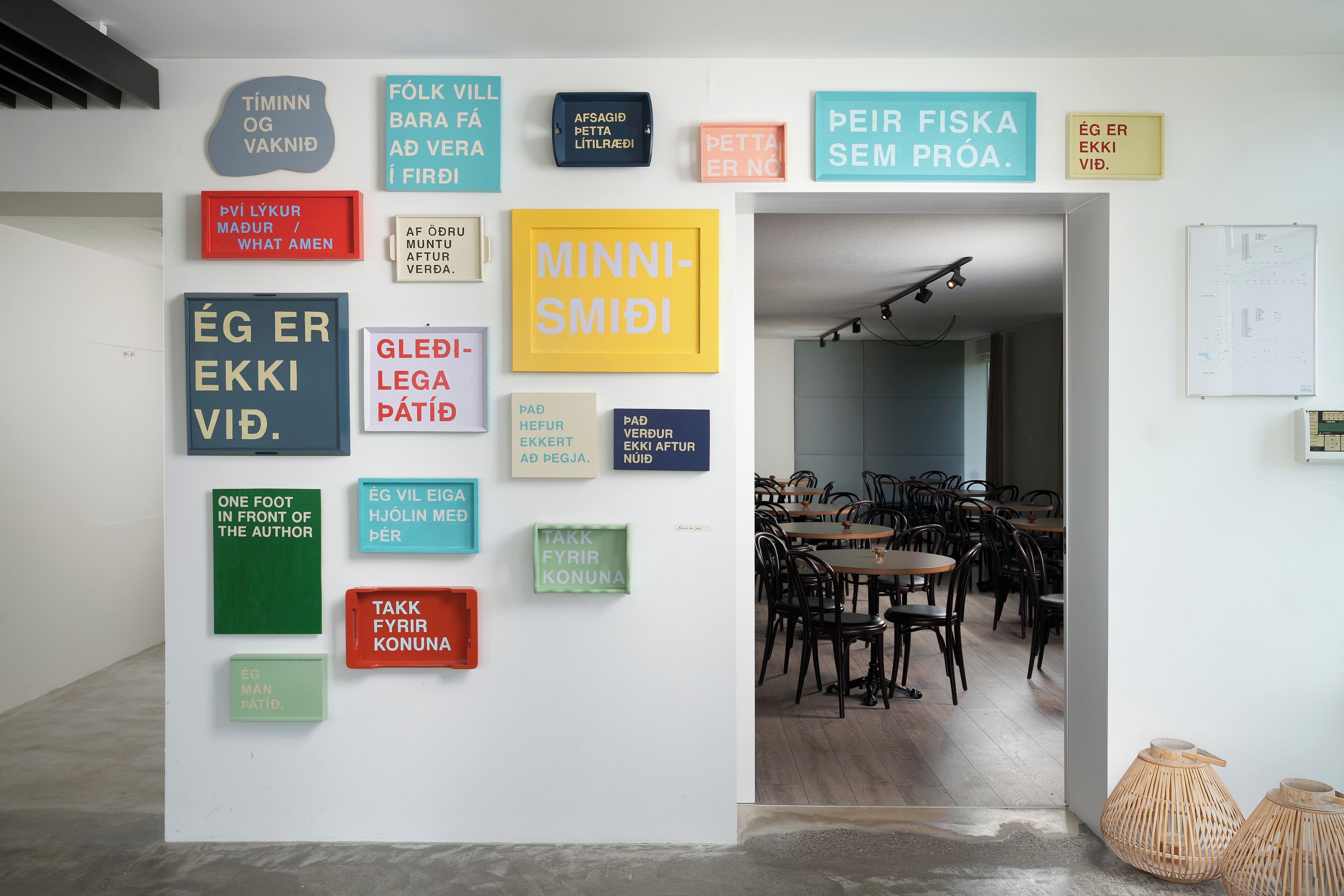 A wall with various framed texts in different colors and languages, a doorway leading to a dining area with tables and chairs.