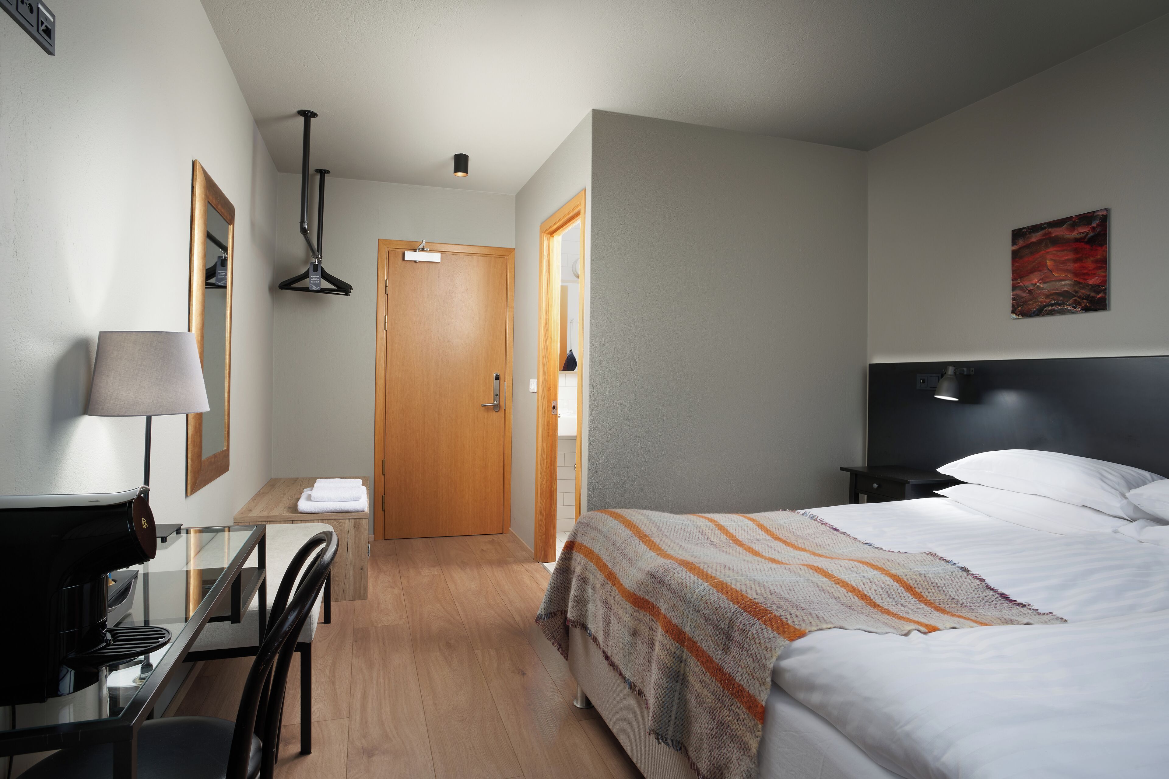 A hotel room with a bed, a desk with a lamp, a chair, a wooden door, and a painting on the wall.