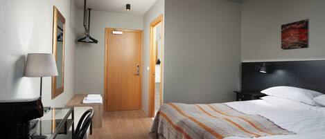 A hotel room with a bed, a desk with a lamp, a chair, a wooden door, and a painting on the wall.
