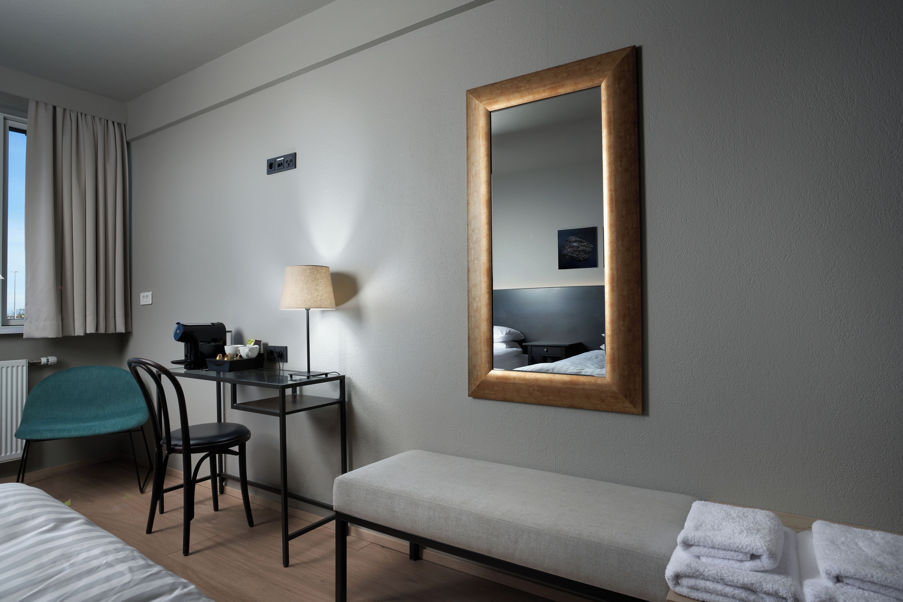 Una camera d'albergo con un letto, una scrivania con lampada, una sedia, una panca e uno specchio grande.