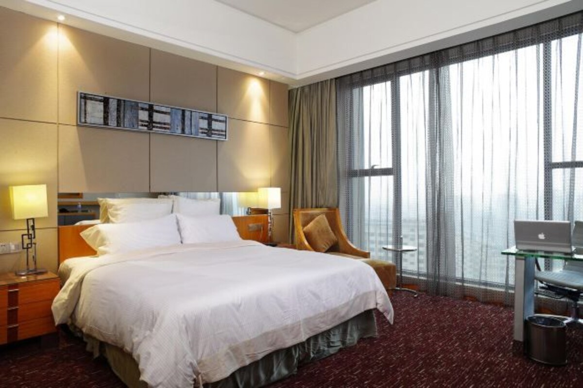 Ein Hotelzimmer mit einem großen Bett, einem Schreibtisch mit einem Laptop, einem Sessel und Blick auf die Stadt durch große Fenster.