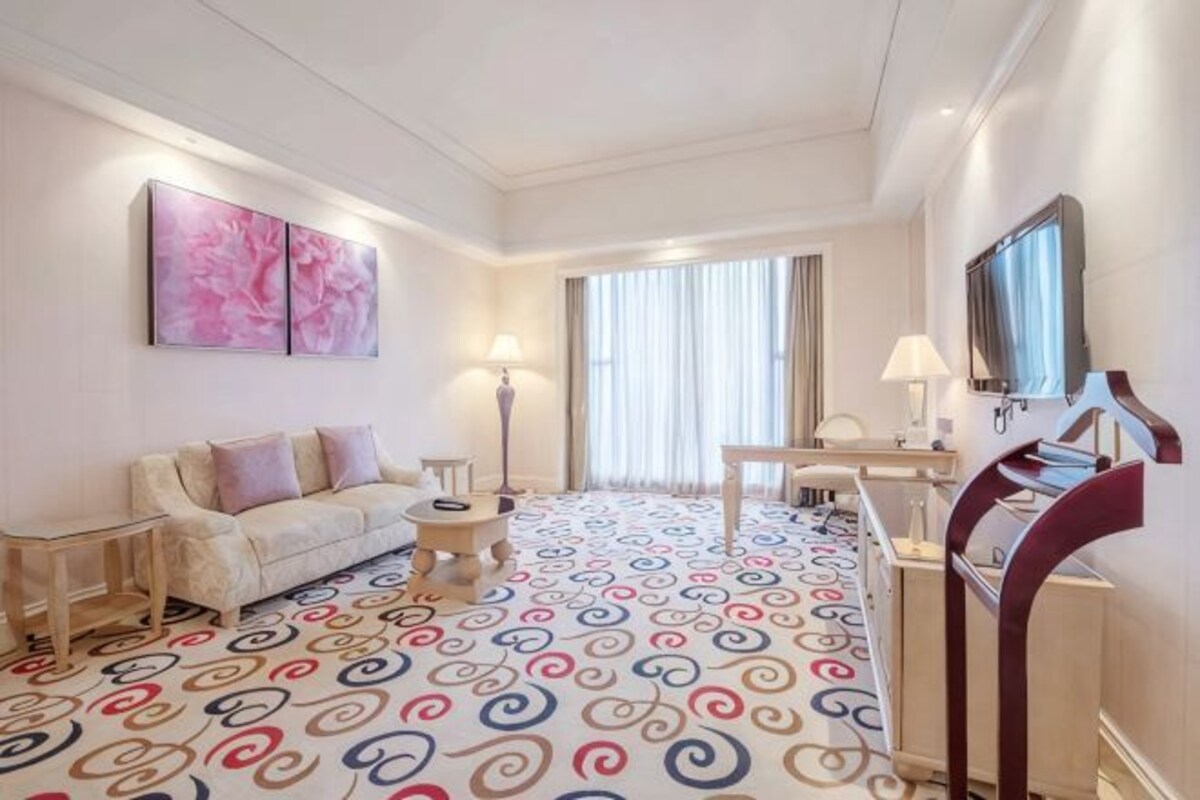 Ein Hotelzimmer mit einem gemusterten Boden, einer Couch, einem Schreibtisch, einem Sessel, einem Fernseher und einem großen Fenster mit Vorhängen.