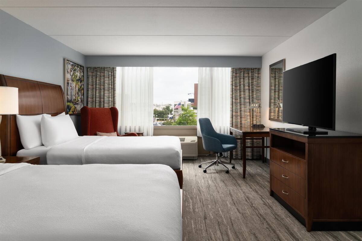 A hotel room with two beds, a desk, a chair, a TV, and a large window with curtains.