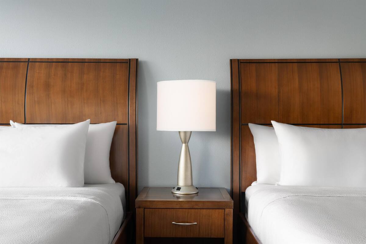 A hotel room with two beds, a nightstand with a lamp, and a wooden headboard.