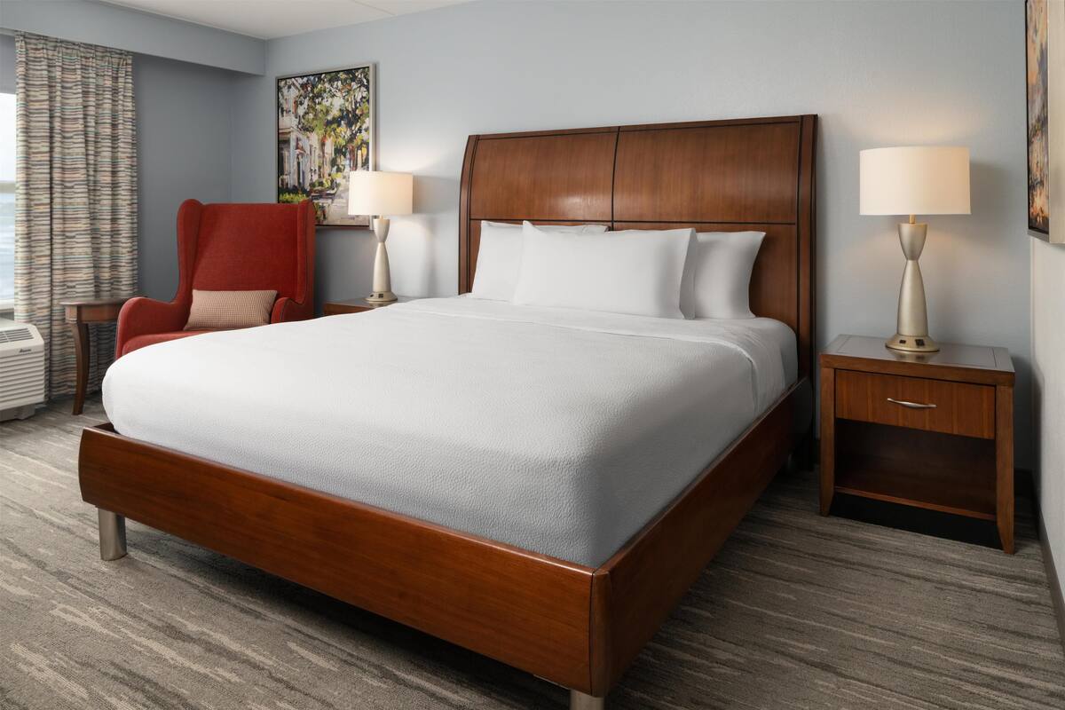 A neatly arranged hotel room with a large bed, a red armchair, two bedside tables with lamps, and a window with curtains.