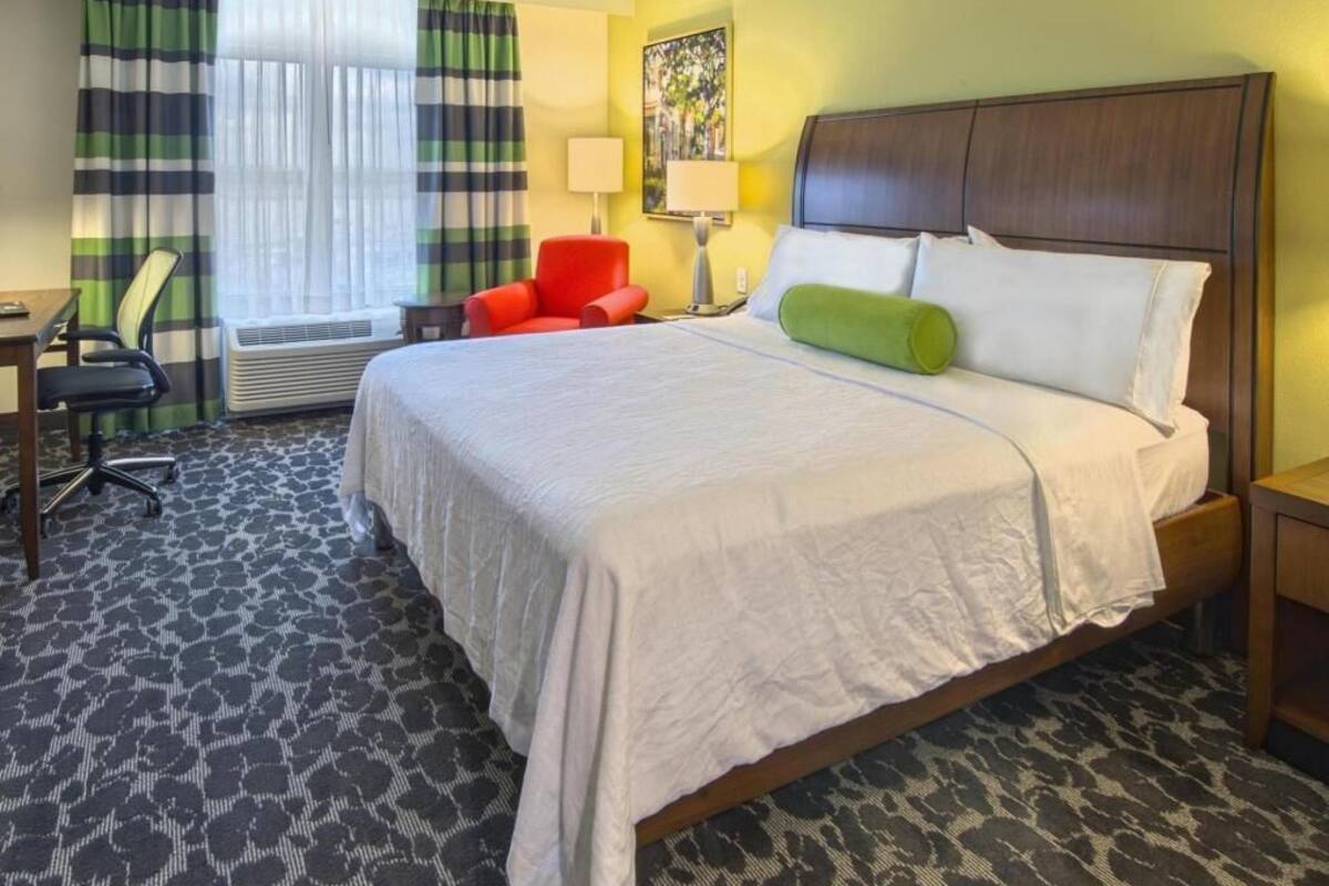 A hotel room with a large bed, a desk, a chair, a lamp, and a painting on the wall.