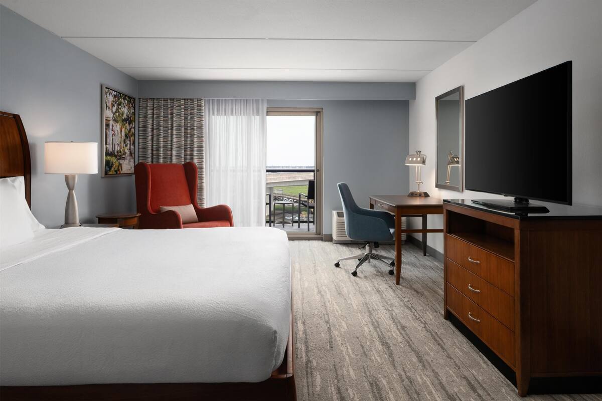 A hotel room with a large bed, a desk, a chair, a TV, and a balcony with a view.