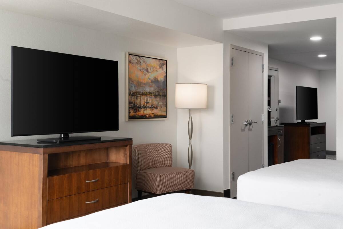 A hotel room with a flat-screen TV, a painting, a lamp, a chair, and a bed.
