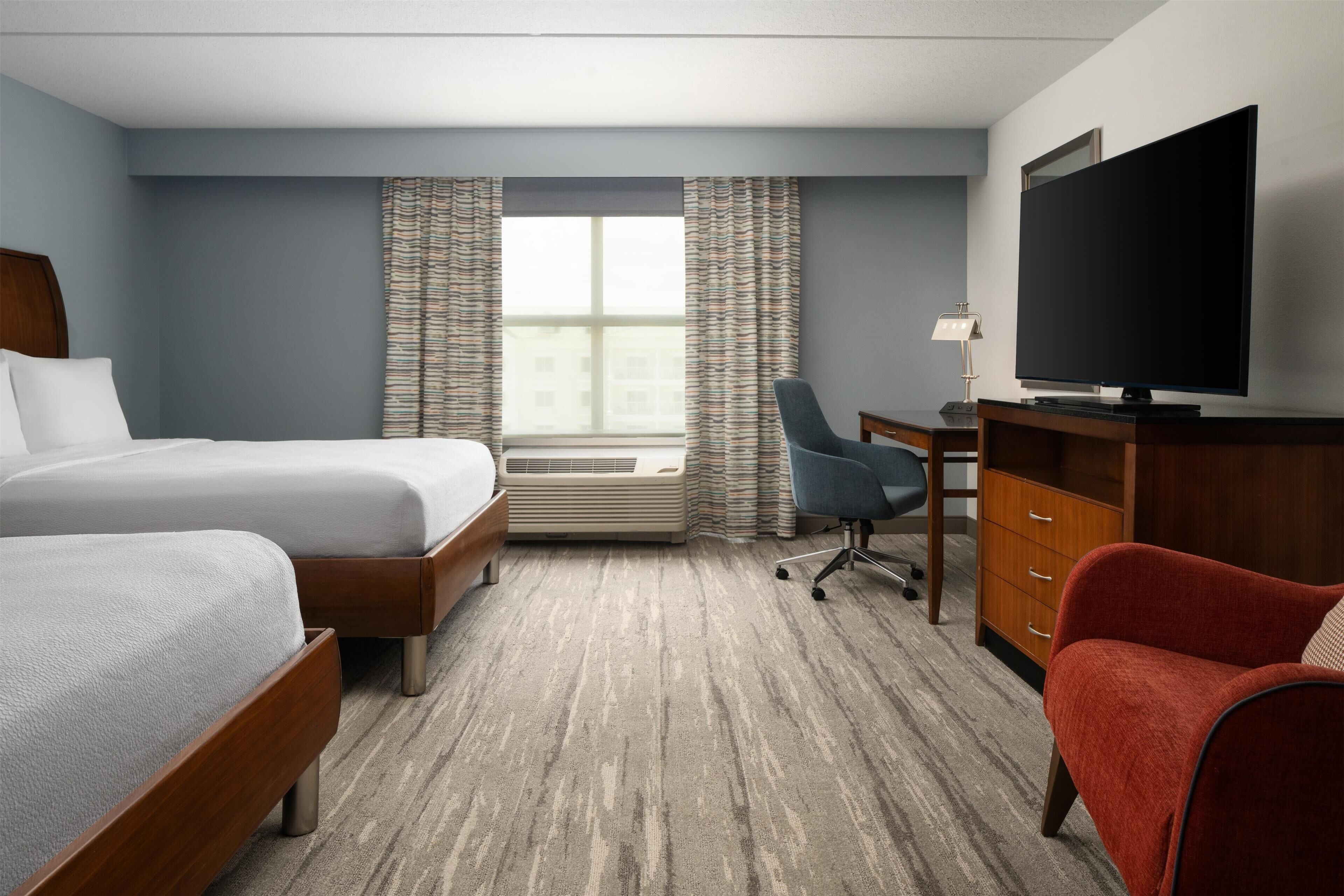 A hotel room with two beds, a desk with a television, a red chair, and a window with curtains.
