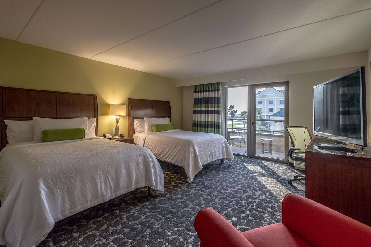 A hotel room with two beds, a desk with a TV, a chair, and a balcony with a view.