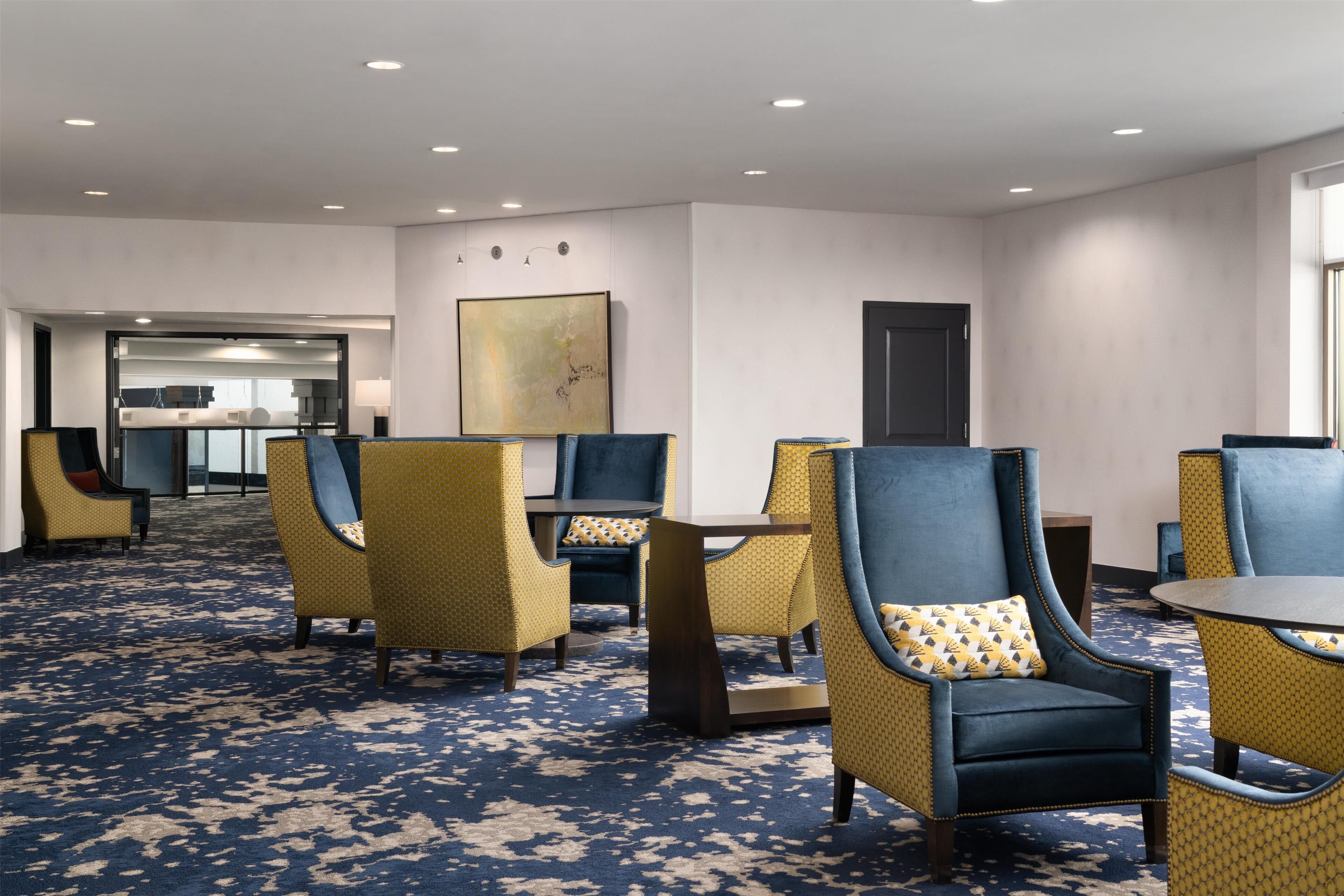 A hotel lobby with a blue patterned carpet, yellow and blue upholstered chairs, a dark wooden coffee table, and a large abstract painting on the wall.