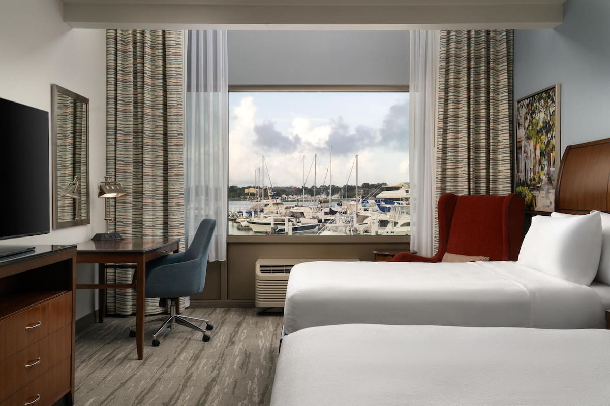 A hotel room with a large window offering a marina view, a desk with a chair, a bed, and a TV.