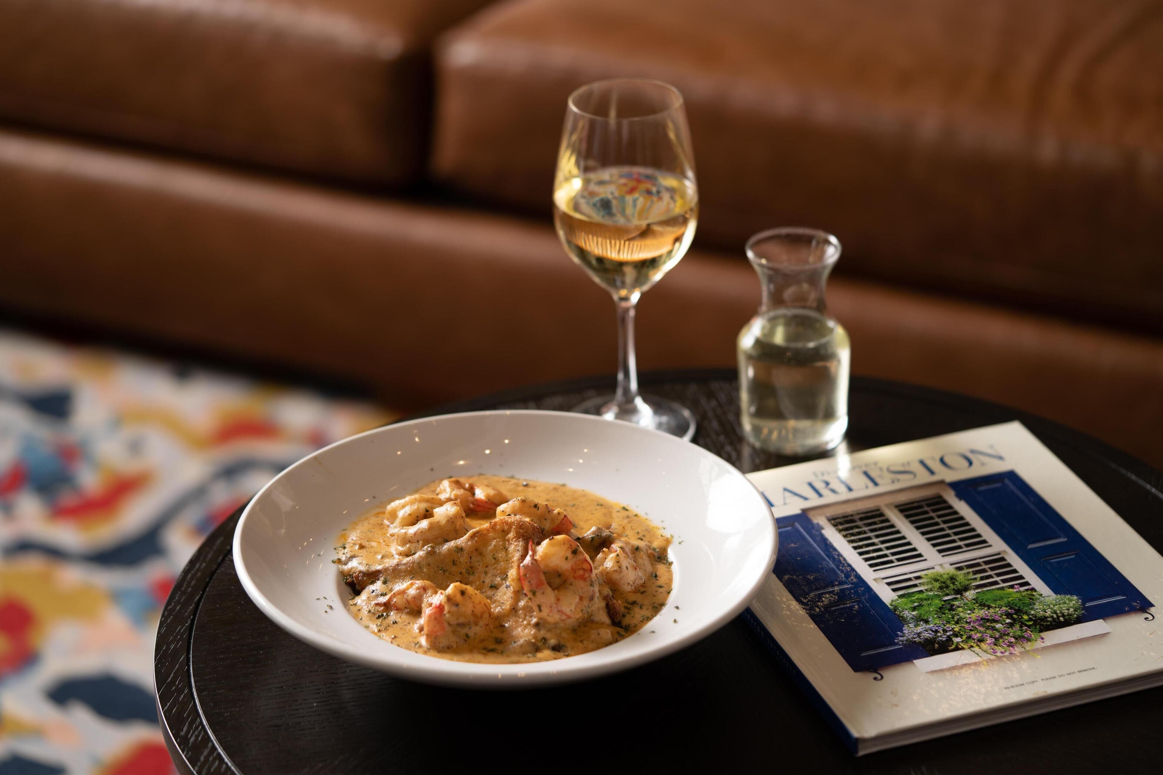 Un plato de camarones con salsa, una copa de vino blanco y una botella de agua sobre una mesa con una revista.