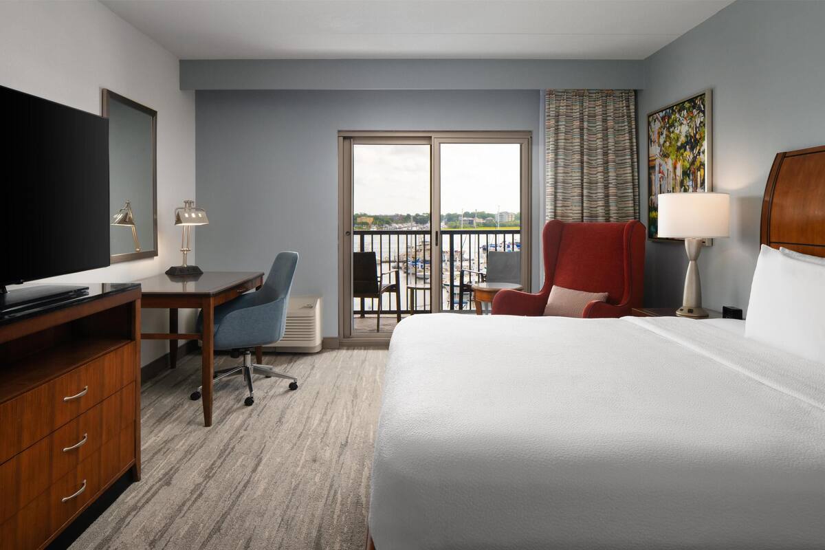 A hotel room with a bed, a desk with a computer, a chair, a TV, and a balcony with a view.