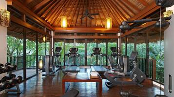 A gym with treadmills, elliptical machines, and free weights, featuring a wooden ceiling and large windows overlooking greenery.