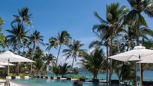 A pool area with lounge chairs and umbrellas, surrounded by palm trees and a clear view of the ocean.