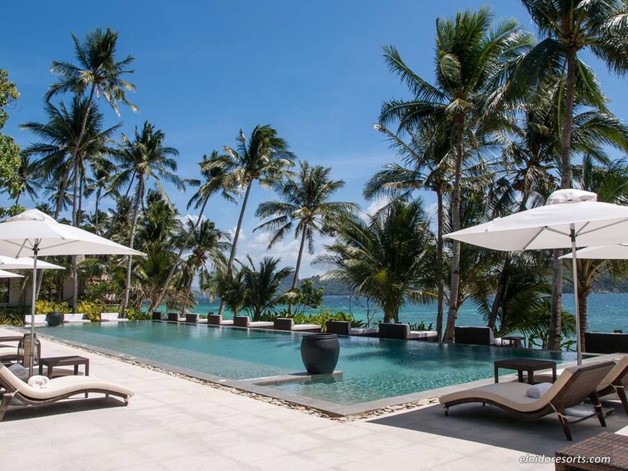 Ein Poolbereich mit Liegestühlen und Sonnenschirmen, umgeben von Palmen und freiem Blick auf den Ozean.