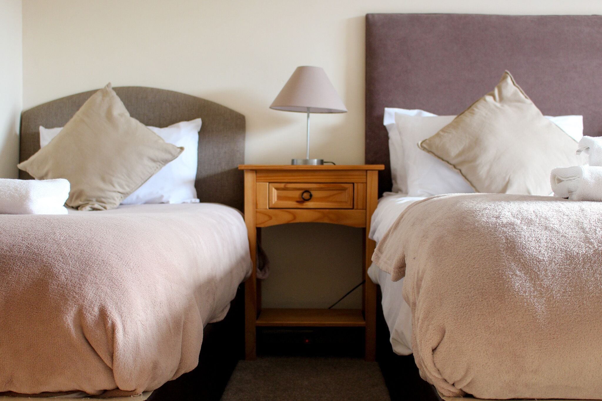 A hotel room with two beds, each with a headboard, a bedside table with a lamp, and a small drawer.