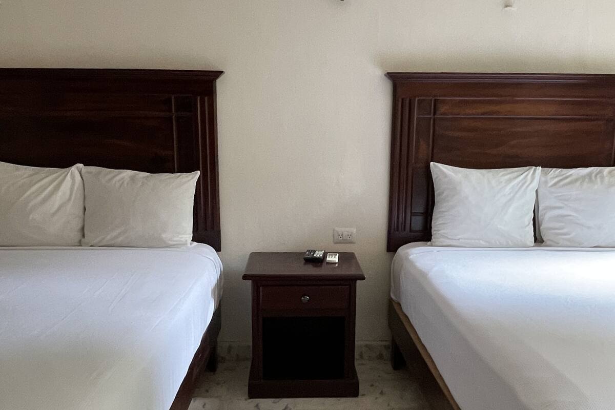Habitación de hotel con dos camas individuales, una mesita de noche de madera y una lámpara de pared.