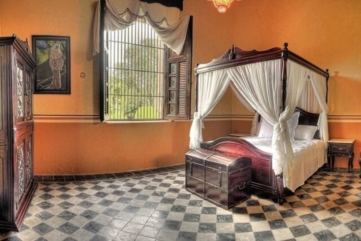 Una habitación con cama con dosel, suelo a cuadros, una ventana con rejas y un cuadro en la pared.