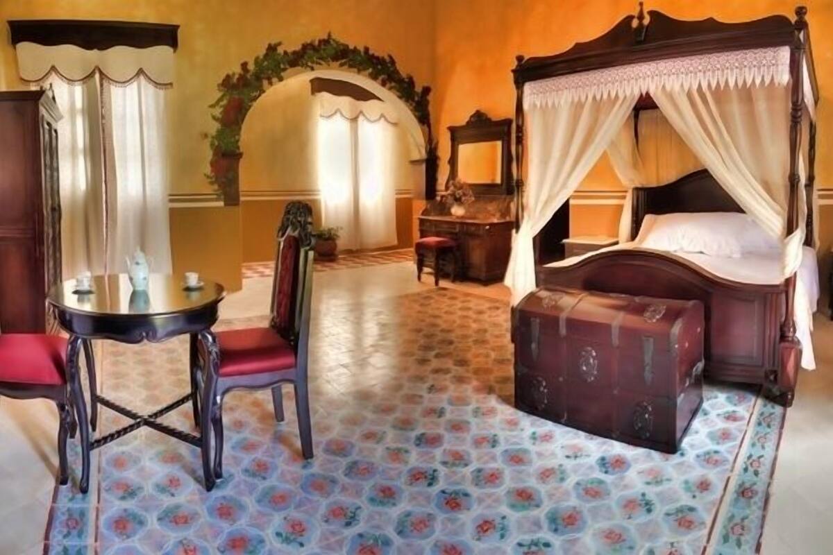 Un dormitorio con una cama con dosel, una zona de estar con una mesa y sillas, y un espejo grande.