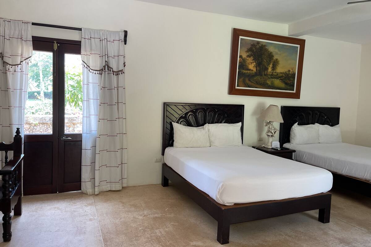 Habitación de hotel con dos camas, un banco de madera, un cuadro en la pared y una ventana con cortinas.