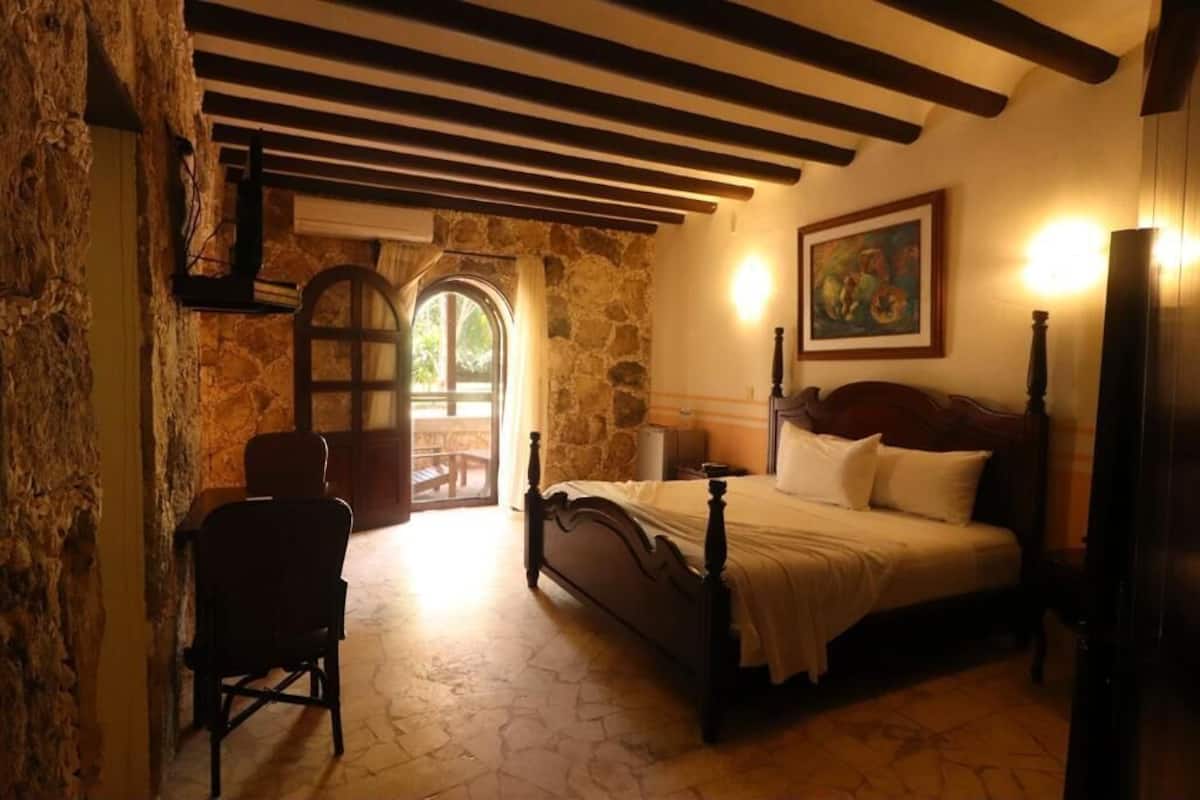 Un dormitorio con pared de piedra, una cama de madera, una silla y un cuadro en la pared.