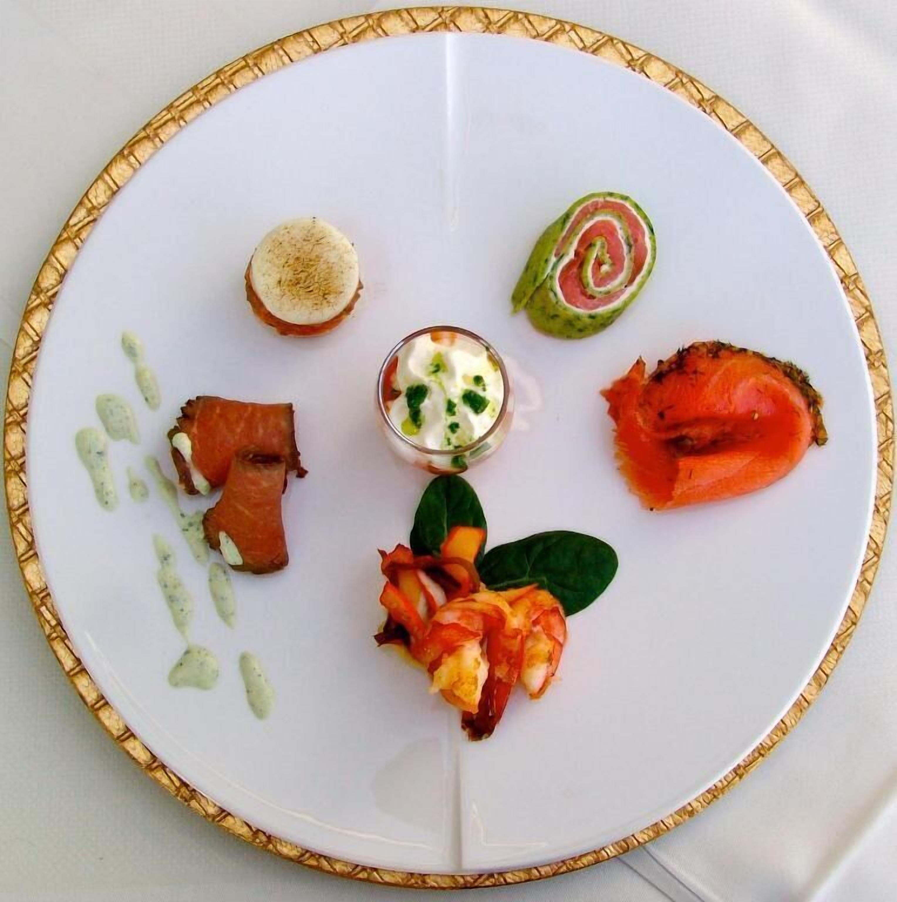 A white plate with a gold rim, containing various food items including smoked salmon, a rolled vegetable, and a small glass of sauce.