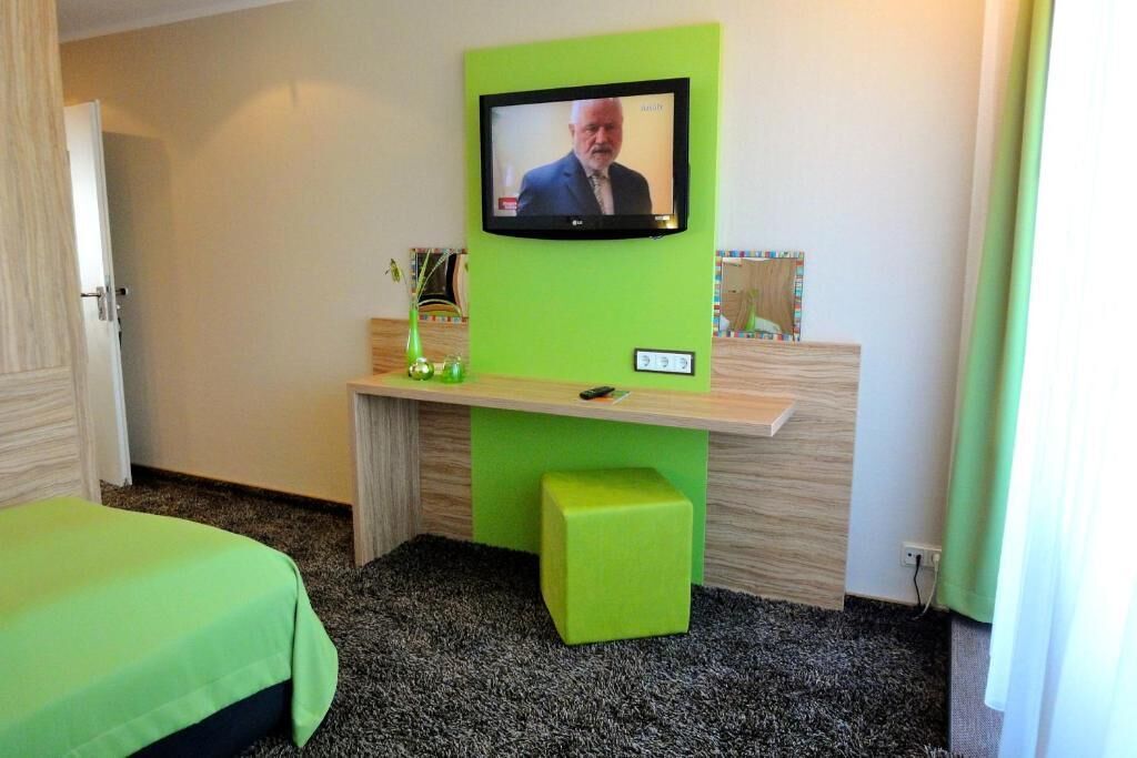 A hotel room with a green wall, a television mounted above a desk, a bed with a green bedspread, and a small green stool.